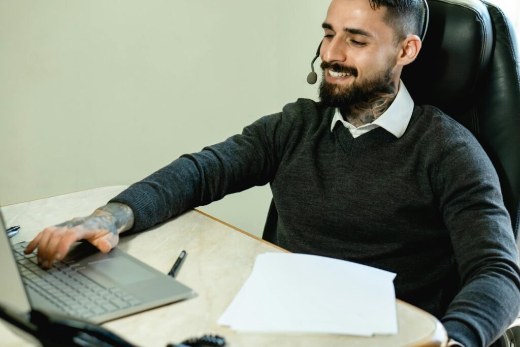 A young tattooed man in a headset works at a desk with a laptop and documents.